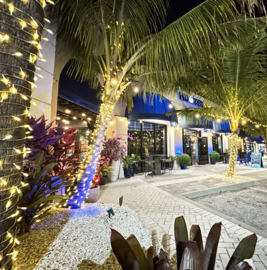 Exterior of Hooked on Jupiter at night, showing the restaurant's entrance with palm trees, string lights, and illuminated signage.